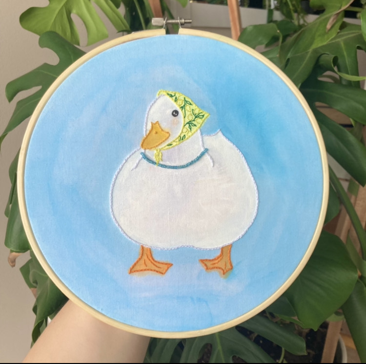 hoop embroidery with blue watercolour background; a very round duck in a lemon-printed bandana and blue pearl necklace